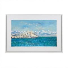 A framed horizontal landscape painting depicting a coastal city with white buildings, blue water, and snow-capped mountains under a blue sky, set in a white frame against a white background.